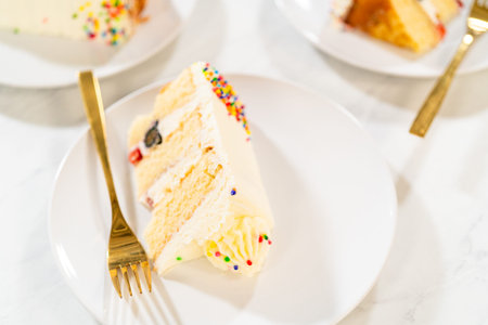 A slice of frosted cake is served on a white plate with a gold fork. Blurred background shows...