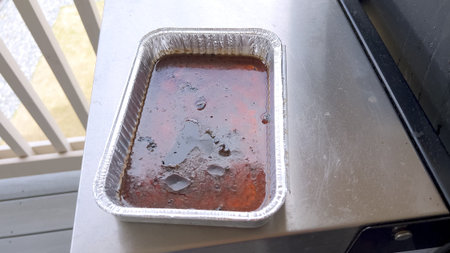 Disposable aluminum tray filled with dark reddish grease and charred food residue sits on a grill...