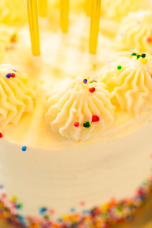 A vertical close-up of a frosted cake shows yellow candles and piped vanilla swirls topped with...