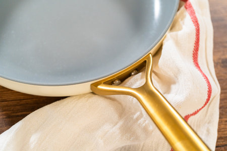 A non-stick frying pan with a gold handle rests on a cream-colored kitchen cloth atop a wooden table. The composition evokes a warm and stylish kitchen aesthetic.の写真素材