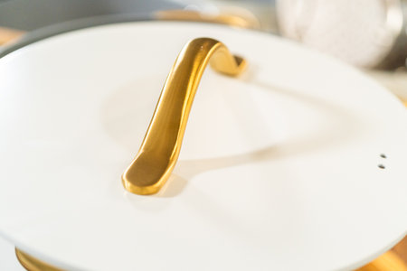 Closeup of a ceramic cookware lid featuring a sculpted gold handle, set against a softly lit kitchen background.の写真素材