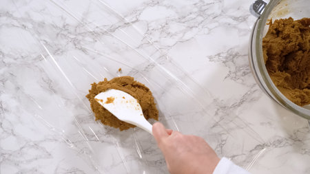A spatula is scooping a portion of brown cookie dough onto a sheet of plastic wrap placed on a marble countertop. The preparation stage is shown under bright lighting in a clean kitchen.の写真素材