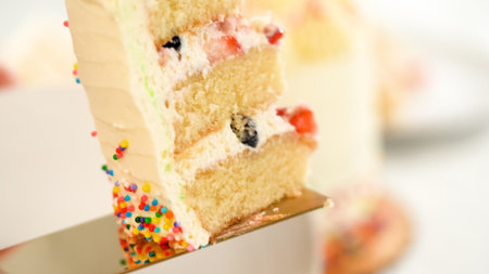 A hand lifts a slice of vanilla cake filled with strawberry and blueberry layers. The inside reveals a moist, fruit-filled interior with colorful sprinkles on the outside.の写真素材