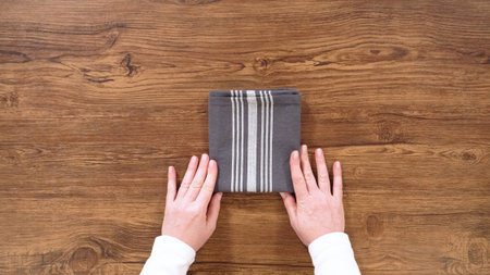 A neatly folded dark striped kitchen towel rests centered on a wooden tabletop. Two hands frame the towel from the bottom, completing the folding process.の写真素材