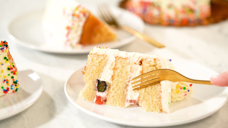 A hand uses a golden fork to cut into a slice of vanilla cake with fruit filling. The cake features...