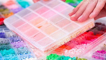 Woman hands gracefully poised over a collection of beads, sorted by color in transparent organizers....