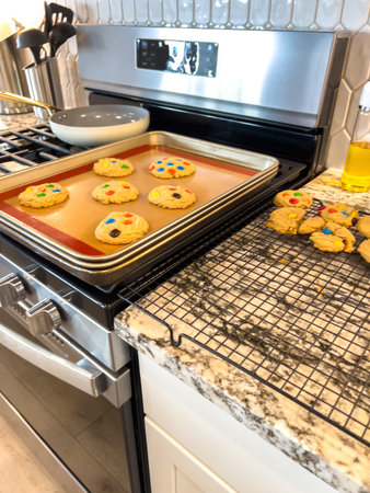Golden brown cookies with colorful candy toppings rest on a wire cooling rack beside a baking tray....