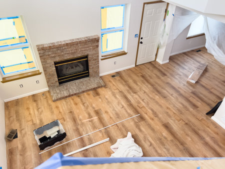 Interior view of a living room under renovation with new laminate wood flooring, tools, and supplies scattered on the floor. The windows and fireplace are partially taped and prepped for painting. Ideal for blogs or articles on home improvement or DIY floのeditorial素材