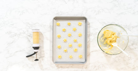 Piped lemon spritz cookie dough on a baking tray next to a bowl of remaining dough. A cookie press...