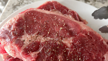 A close-up view of a raw ribeye steak with seasoning on top, placed on a white plate. The marbling of the meat is clearly visible, ready for grilling.の写真素材