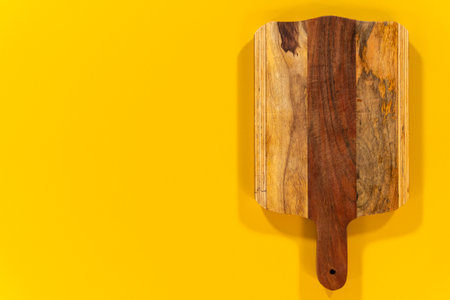 A wooden cutting board with a handle is centered on a vibrant yellow background. The image highlights the contrasting wood tones and the handmade appearance of the board.の写真素材
