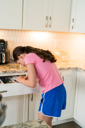 A young girl in a pink shirt and blue shorts is leaning over a kitchen counter, inspecting freshly...
