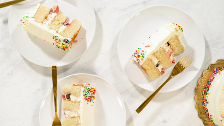 Three slices of vanilla celebration cake with colorful sprinkles are arranged on white plates. Gold...