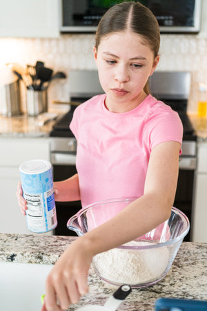 A girl in a pink shirt prepares dry ingredients in a glass bowl, holding a container of baking...