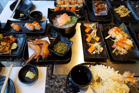 Variety of food with fried chicken wings, grilled bacon wrapped golden needle mushroom, Crab stick salad, sushi and sashimi on dark background in restaurant, food of hotel buffet, ready to eatの写真素材