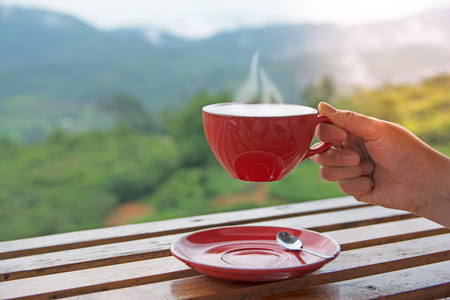 Hand holding red cup of hot coffee with smoke with green natural mountain as background.の写真素材