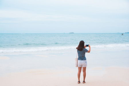 A young woman holding mobile phone to shoot the photo at a beautiful beach and white sand.の写真素材