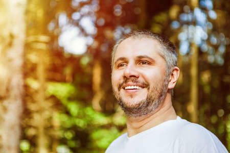 Portrait of a smiling surprised man with beard standing and looking ahead against a green trees in sunny forestの写真素材