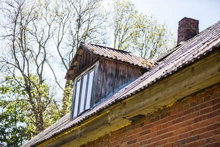 Old wooden roof with small window and chimney in rural Latvia, Europe. Red brick wall weathered with texture. Trees in background.の写真素材