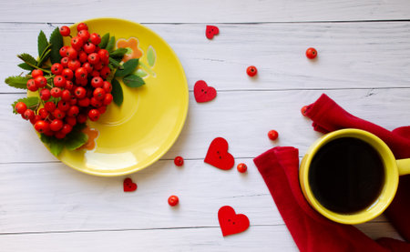 Yellow mug and saucer on white wooden background flat lay. Ripe rowan berries lie on saucer, red...