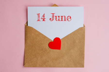 Kraft envelope with a white sheet of paper and a date 14 june, with red heart. Flat lay on pink...