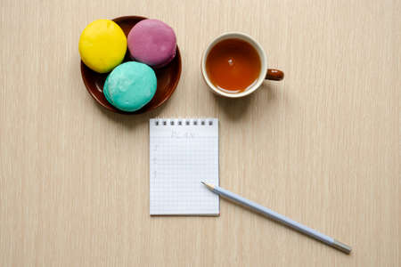 A mug with tea and macaroons on saucer on a wooden table, flat lay. Notebook with pencil, business...