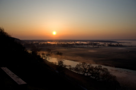 Picturesque fairytale sunrise over a foggy meadow and river on a summer morning. Empty bench on the mountain overlooking the field. World Environment Day Concept.の写真素材