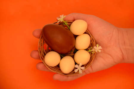 Chocolate eggs in a basket with flowers in hand flat lay. On an orange background Easter eggs, milk...
