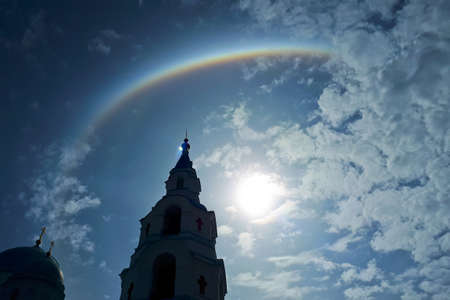 Rainbow halo in the blue sky with white clouds. Magical natural rainbow effect with the sun. Temple silhouette against the beautiful skyの写真素材