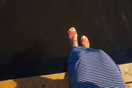 Woman's legs in red sandals over dark water of the lake on a sunny dayの写真素材