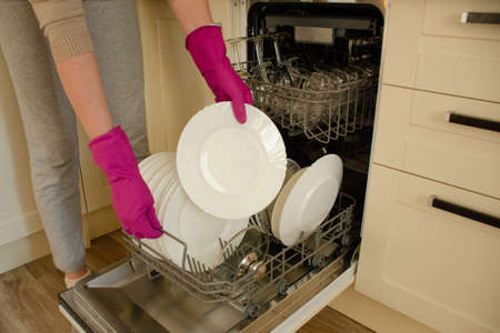 Housewife in pink rubber gloves takes out clean dishes from the dishwasher in the kitchenの写真素材