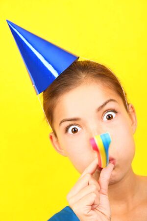 Funny party woman in blue on yellow background. Beautiful mixed race / asian caucasian model with party hat blowing a party blower at the camera. の写真素材