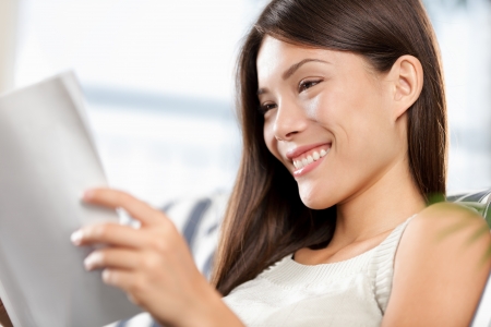 mixed race chinese asian / caucasian woman reading magazine relaxing sitting in sofa at home. Model smiling happy.の写真素材