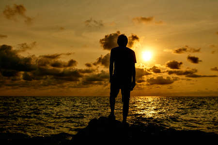 Silhouette of a man standing on a rock by the sea at sunsetの写真素材