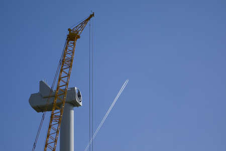 An passenger aircraft is drawing contrails behind a wind turbine under constructionの写真素材
