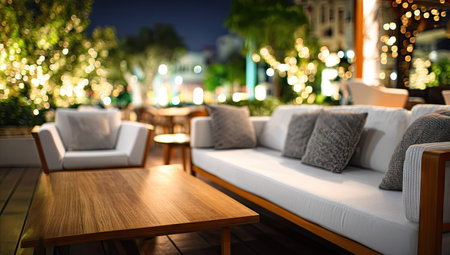 An outdoor lounge area at night features comfortable seating with a wooden table and ambient lighting. The scene displays a modern design with neutral colors and textures, with blurred background elements suggesting a social gathering. This image could be used for commercial projects focusing on lifestyle or interior design.の素材