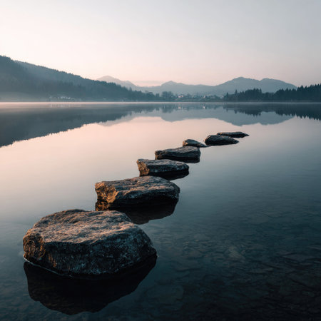 A line of stepping stones provides a pathway across a calm lake, leading towards distant hills. The scene features soft light, creating a serene atmosphere. The color palette includes muted tones and reflections in the water. This image can be used for a variety of commercial and editorial projects.の素材