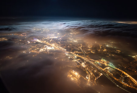 An aerial photograph captures a nighttime cityscape, with glowing lights illuminating roads and structures. A layer of clouds partially obscures the view, adding depth and texture to the image. The composition exhibits a dramatic contrast of light and shadow, suitable for various commercial and editorial applications.の素材