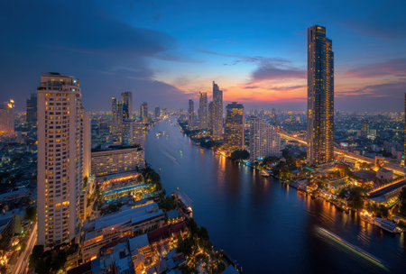 An elevated view captures a cityscape at twilight with illuminated skyscrapers. The scene displays a river winding through the urban environment, reflecting ambient light. The composition highlights architectural elements, showcasing a blend of lights and shadows. This image could serve various uses, from commercial projects to editorial illustrations.の素材