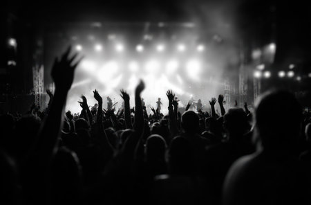 A crowd of people raise their hands in silhouette towards bright stage lights. The monochrome image highlights the contrast between dark figures and a glowing background. The composition suggests an indoor venue, likely during a performance. This image could be used for various editorial or commercial purposes.の素材