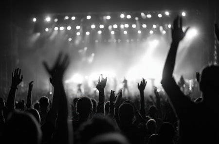 A crowd of people raise their hands in front of a brightly lit stage in a monochrome photograph. The image features a shallow depth of field, with the audience silhouetted against the bright lights. This image could be used for promoting music events, entertainment news, or artistic projects.の素材