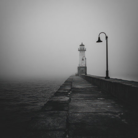 A lighthouse stands at the end of a stone pier shrouded in dense fog. The monochromatic image features strong horizontal lines and muted tones, creating a sense of solitude. The composition utilizes symmetry, with a solitary light post adding depth. Suitable for various editorial and commercial applications.の素材