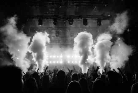 This monochrome image captures a concert stage enveloped in billowing clouds of smoke. The scene features bright stage lights illuminating the smoke, creating a dramatic atmosphere. Silhouetted figures of the audience are seen with raised hands, suggesting an energetic performance. Suitable for editorial and commercial use.の素材