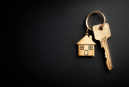 A golden key with a house-shaped charm is displayed prominently against a dark background. The composition features a close-up shot with a shallow depth of field, emphasizing the key's details. The lighting is focused, casting soft shadows. The image is suitable for various commercial purposes, including property and finance.の素材