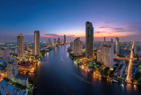 An aerial view presents a city skyline at dusk, highlighting numerous tall buildings bordering a wide river. The scene showcases modern architectural designs and illuminated structures. The image's color palette combines dark blue, orange and yellow hues, suggesting a nighttime environment. This image could be used for various commercial or editorial projects.の素材