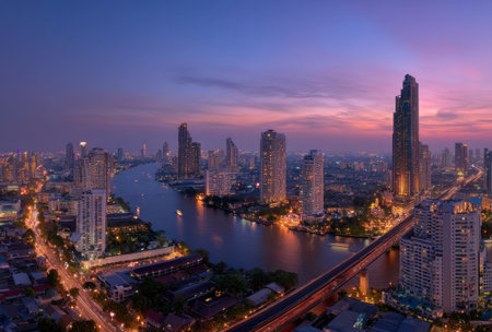 An elevated view captures a cityscape at twilight, highlighting illuminated skyscrapers along a river. The composition showcases a balance of architectural elements and natural features under a colorful sky. This image is suitable for various commercial purposes, including editorial content and promotional materials.の素材