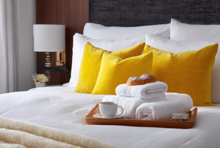 A well-lit hotel bedroom presents a comfortable arrangement. Focus is on a white bed with bright yellow pillows and a wooden tray. The tray holds a cup, towels, and pastries. The composition features a bedside lamp and neutral colors, suitable for diverse commercial applications.の素材