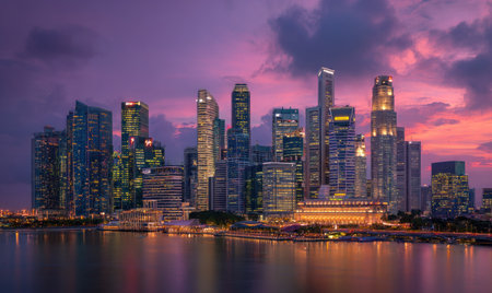 A cityscape is illuminated at dusk. High-rise buildings are silhouetted against a colorful sky. The composition emphasizes architectural forms reflected on the water. The scene shows the interplay of light and shadow, potentially suitable for commercial and illustrative purposes.の素材