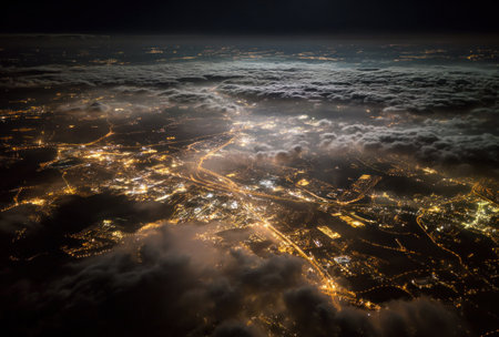 An aerial perspective showcases a sprawling city illuminated by artificial lights at night. Clouds partially obscure the land, creating a dramatic interplay of light and shadow. The image's composition highlights the urban landscape under a darkened sky. This scene may be useful for various commercial and editorial applications.の素材