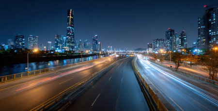 An elevated perspective captures a highway at night, showcasing blurred light trails from moving vehicles. The scene is bathed in cool tones, with illuminated buildings outlining the cityscape. The composition emphasizes a sense of movement and urban atmosphere, suitable for various commercial or editorial applications.の素材